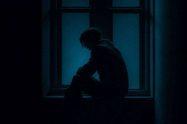A lone figure sitting in the dark with bowed head, isolated by a window