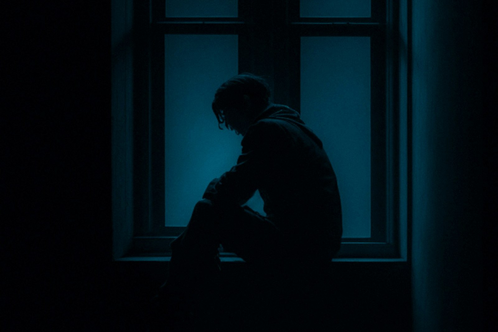 A lone figure sitting in the dark with bowed head, isolated by a window