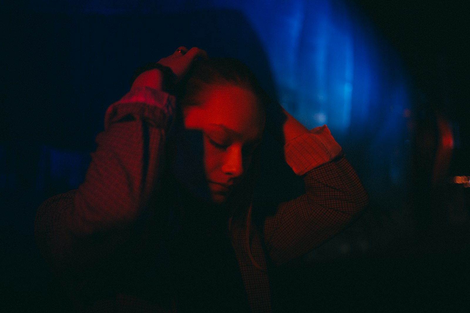 A young person with bowed head and hands on their head in dim light