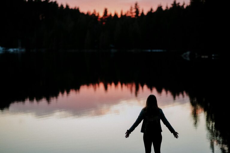 A person standing at the edge of a still lake at sunset with arms open